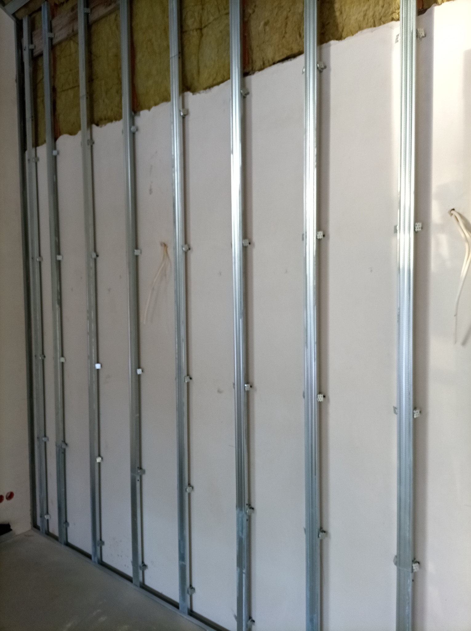 Montaż płyty gipsowo-kartonowej na metalowym stelażu z widoczną izolacją z wełny mineralnej. Ściana w trakcie budowy, widoczne elementy mocujące.