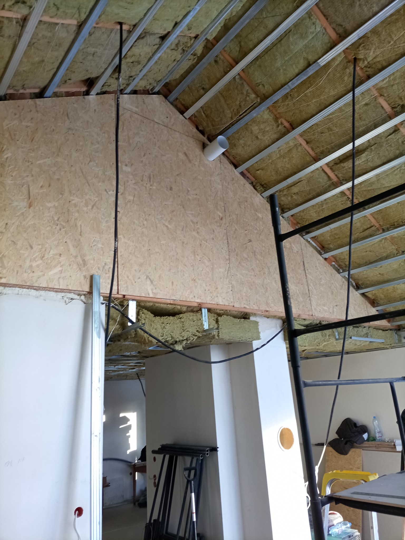 Skośny sufit z izolacją z wełny mineralnej i płyt OSB, widoczne profile metalowe. Fragment ściany z profilem. W tle widoczne narzędzia i elementy rusztowania.