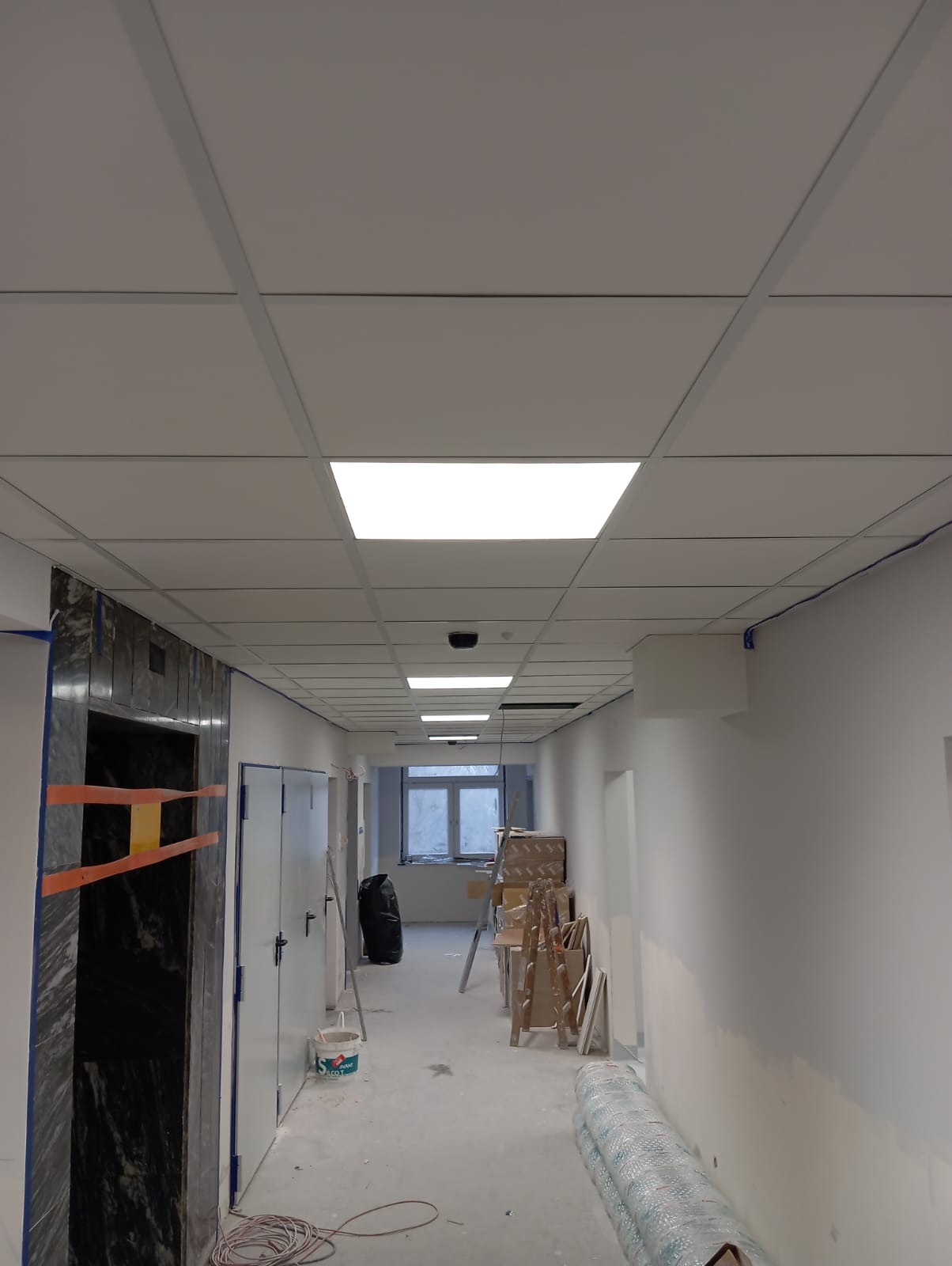 Korytarz z podwieszanym sufitem kasetonowym i oświetleniem LED. Widoczne materiały budowlane i drabina. Świeżo pomalowane ściany.