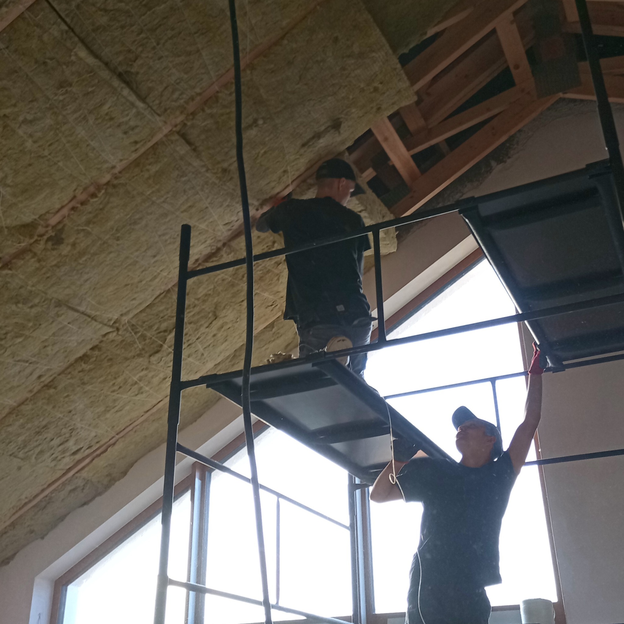 Montaż izolacji poddasza z rusztowania. Dwóch pracowników w trakcie pracy, widoczne okna dachowe i drewniana konstrukcja dachu. Ocieplenie z wełny mineralnej.