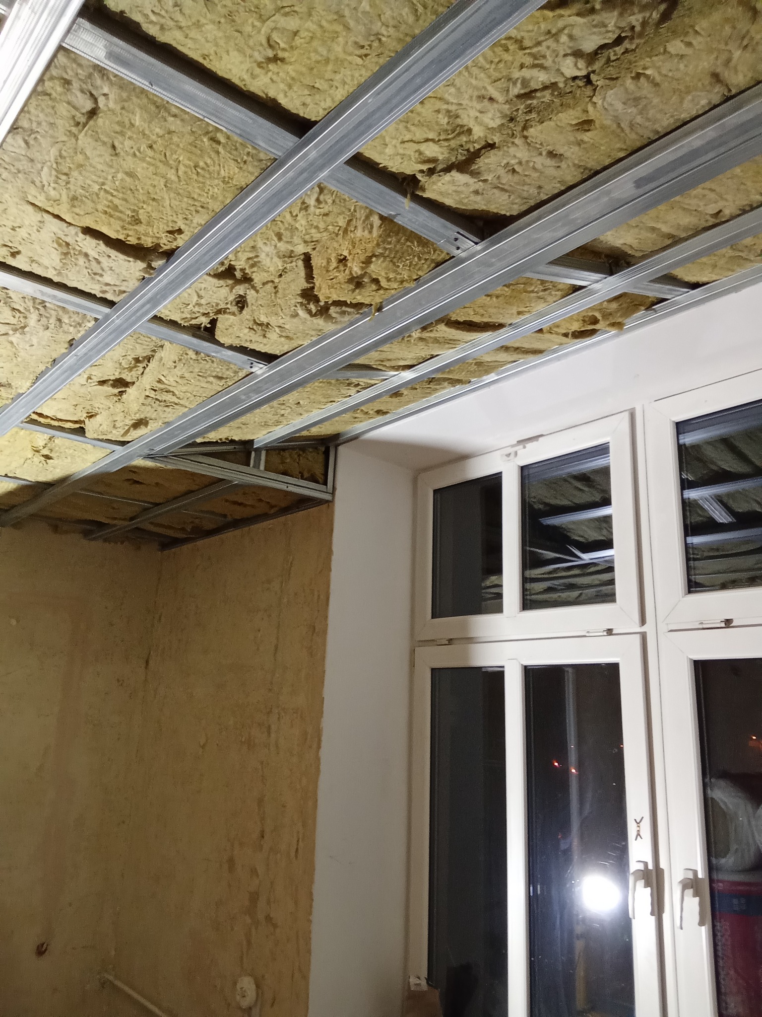 Montaż izolacji z wełny mineralnej w metalowym stelażu sufitu podwieszanego, widoczne okno i fragment ściany w trakcie prac wykończeniowych.