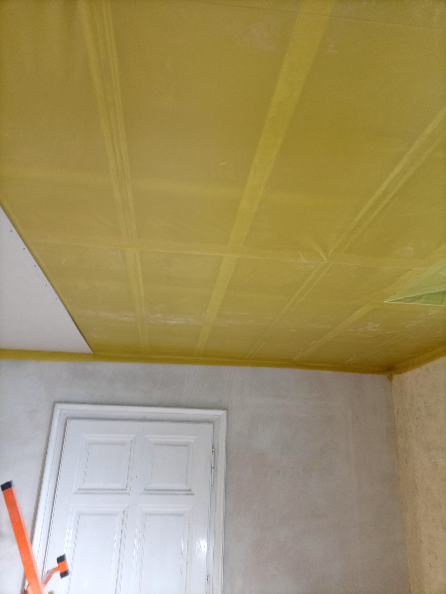 Żółta folia ochronna na suficie z płyt karton-gips, zabezpieczająca przed malowaniem. Widoczna biała stolarka drzwiowa i fragment drabiny malarskiej.