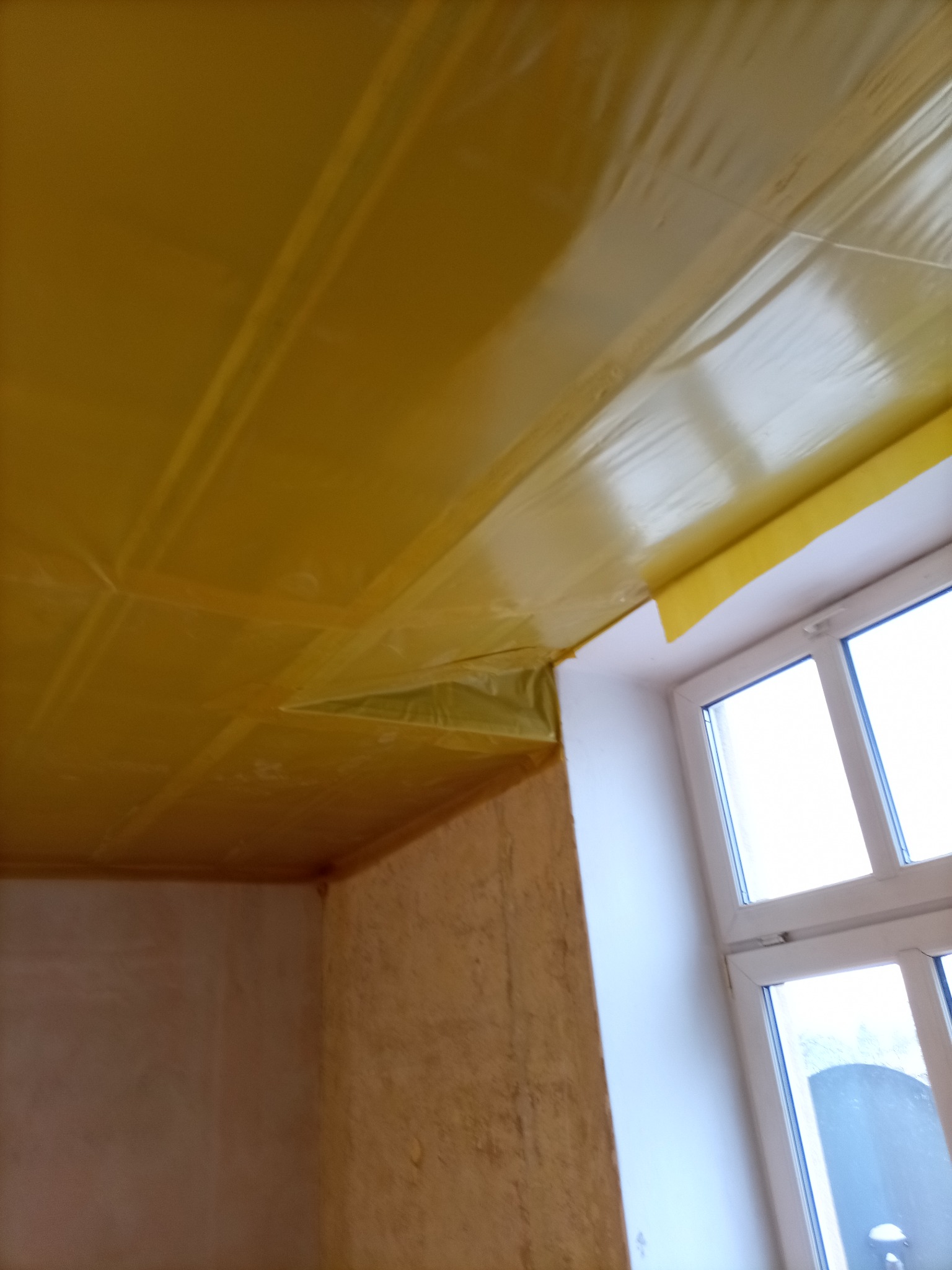 Pokrycie sufitu folią ochronną przed pracami wykończeniowymi. Widoczny fragment okna i ściany z nierówną powierzchnią. Przygotowanie do malowania lub gładzi.