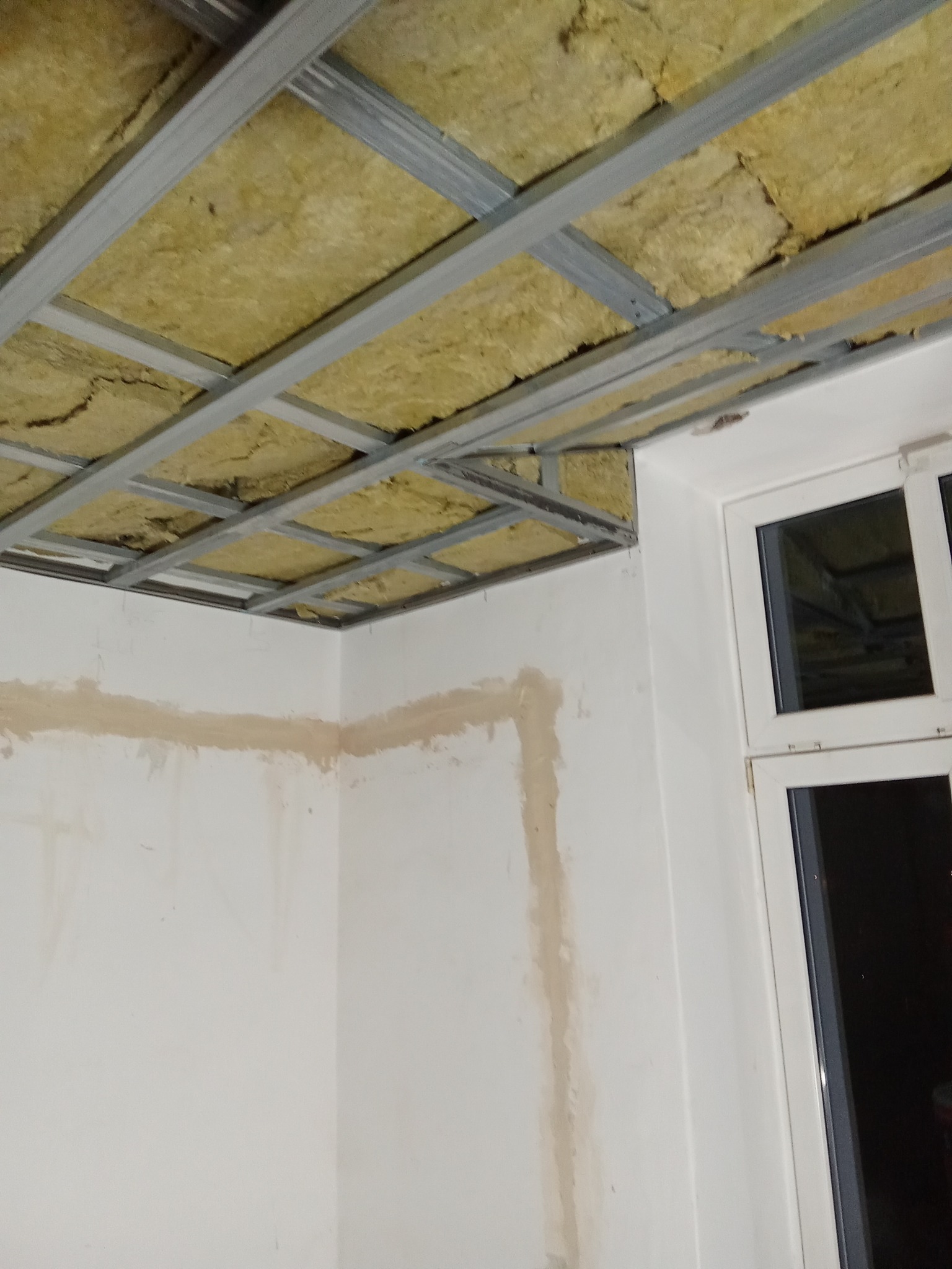 Montaż sufitu podwieszanego z izolacją z wełny mineralnej. Widoczne profile metalowe i fragment ściany z śladami po gładzi.