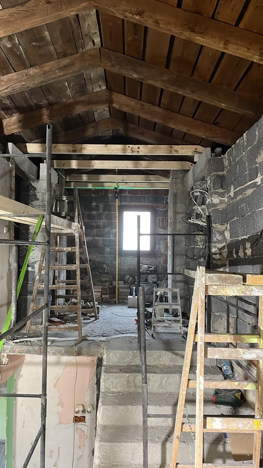 Wnętrze w trakcie remontu poddasza: surowe ściany z cegły, drewniana konstrukcja dachu, schody betonowe i drabiny. Widoczne instalacje i narzędzia. Oświetlenie naturalne z okna.