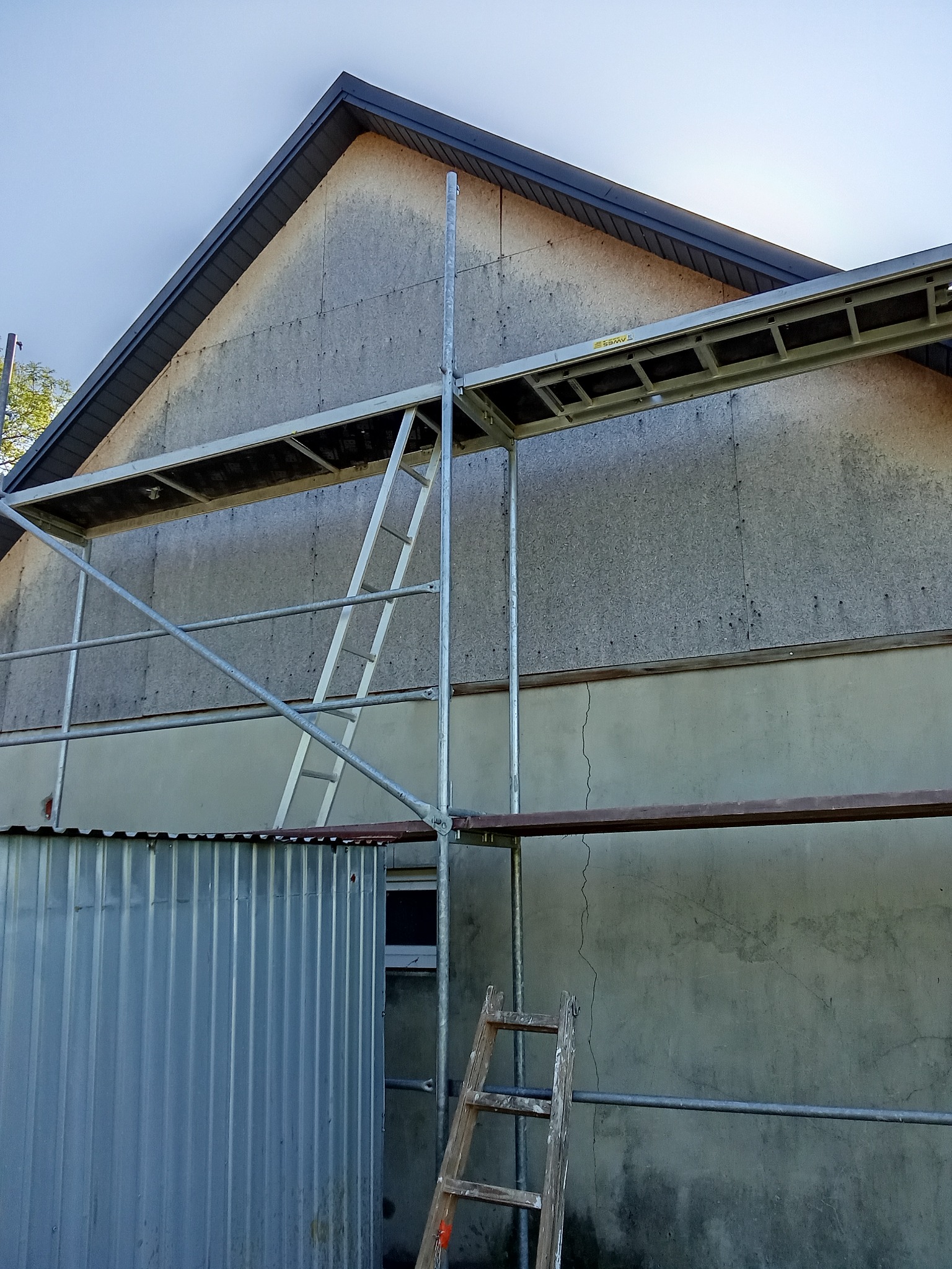 Elewacja domu w trakcie prac z rusztowaniem. Widoczna płyta OSB na szczycie i fragment blaszanej konstrukcji. Ujęcie z dołu, w tle błękitne niebo.