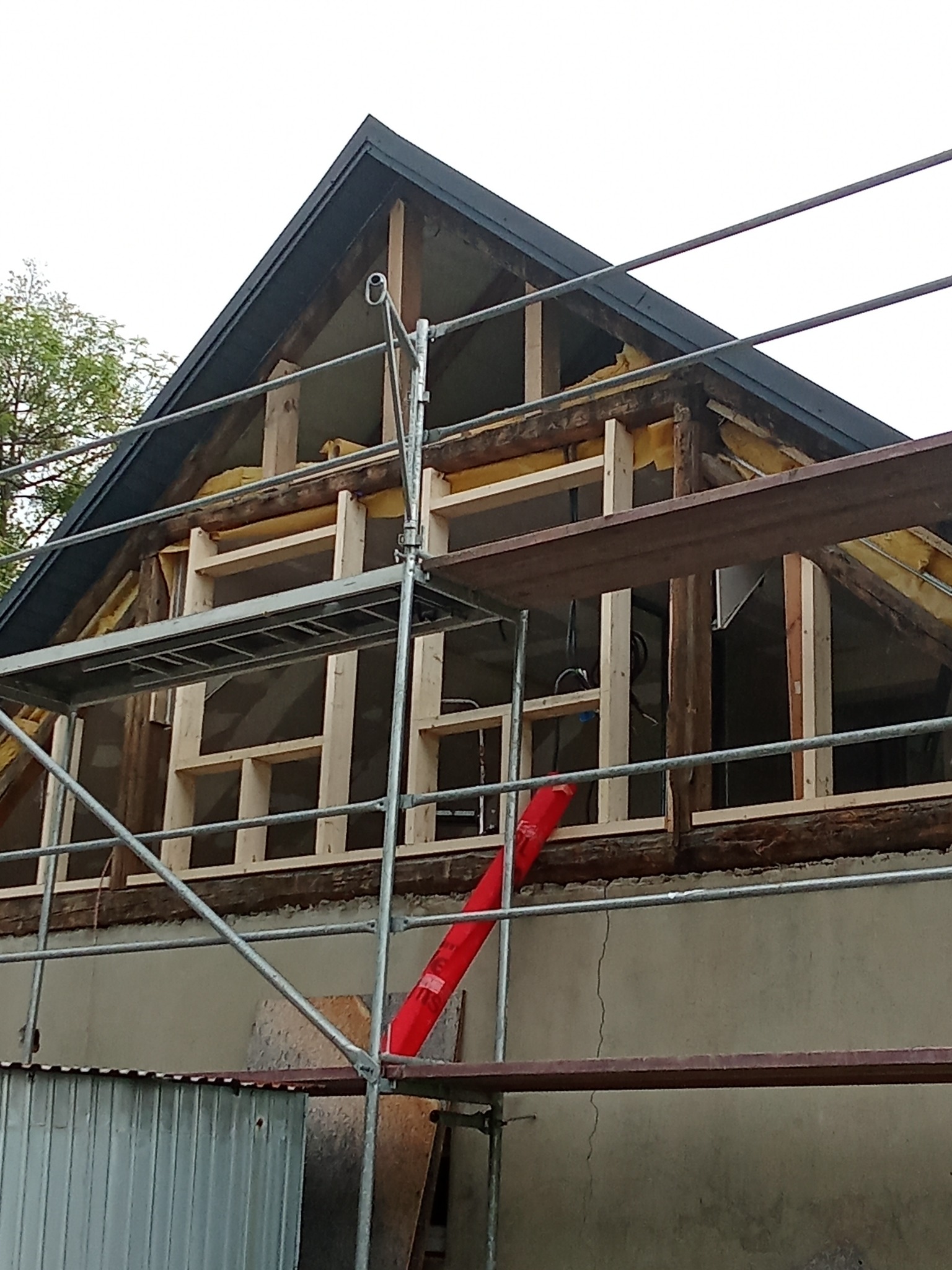 Konstrukcja dachu w trakcie przebudowy, widoczne drewniane elementy, izolacja i rusztowanie. Prace remontowe na zewnątrz budynku.
