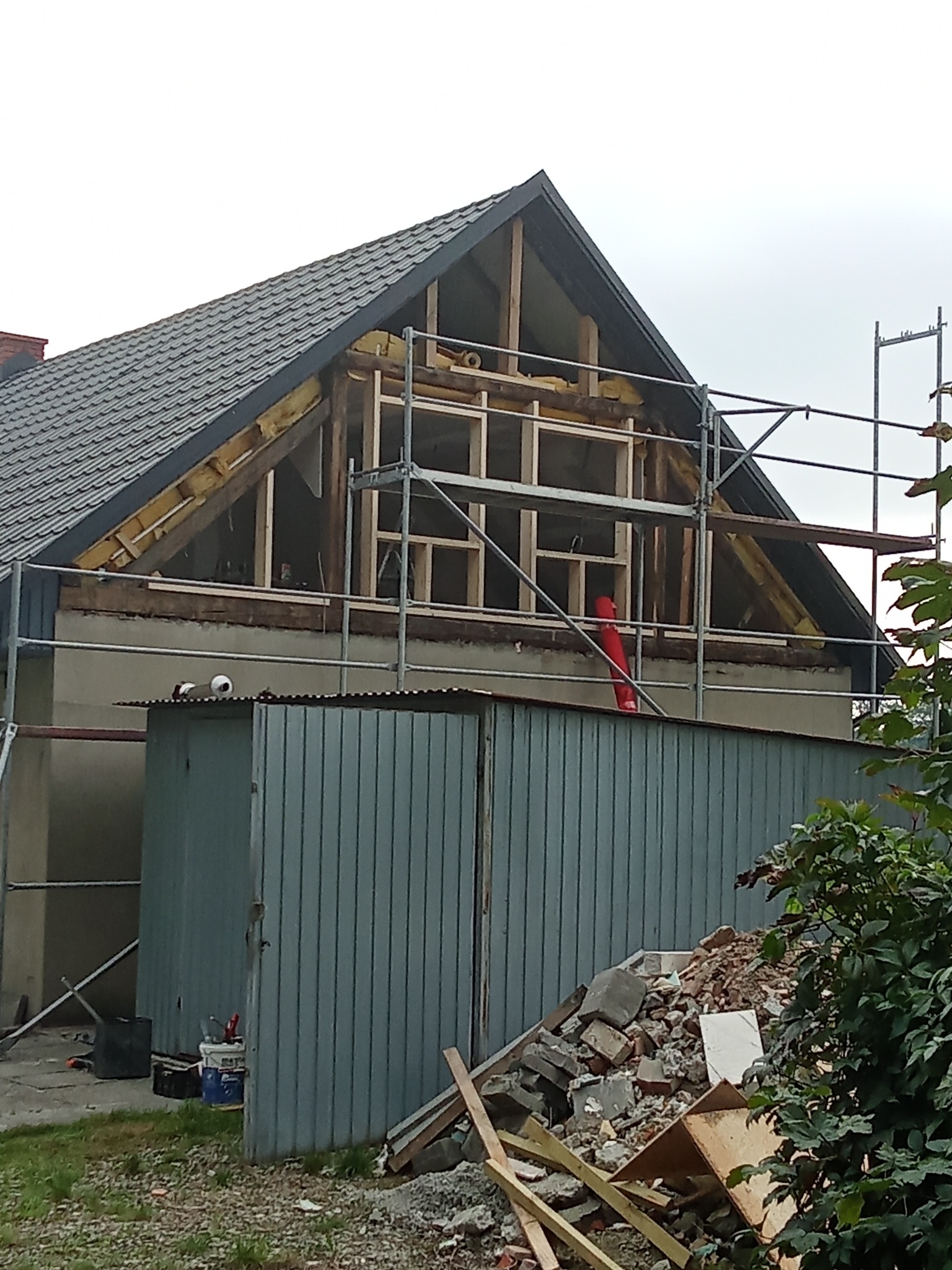 Dom w trakcie remontu poddasza z widocznymi rusztowaniami, drewnianą konstrukcją i izolacją. Na pierwszym planie blaszany garaż i gruz.