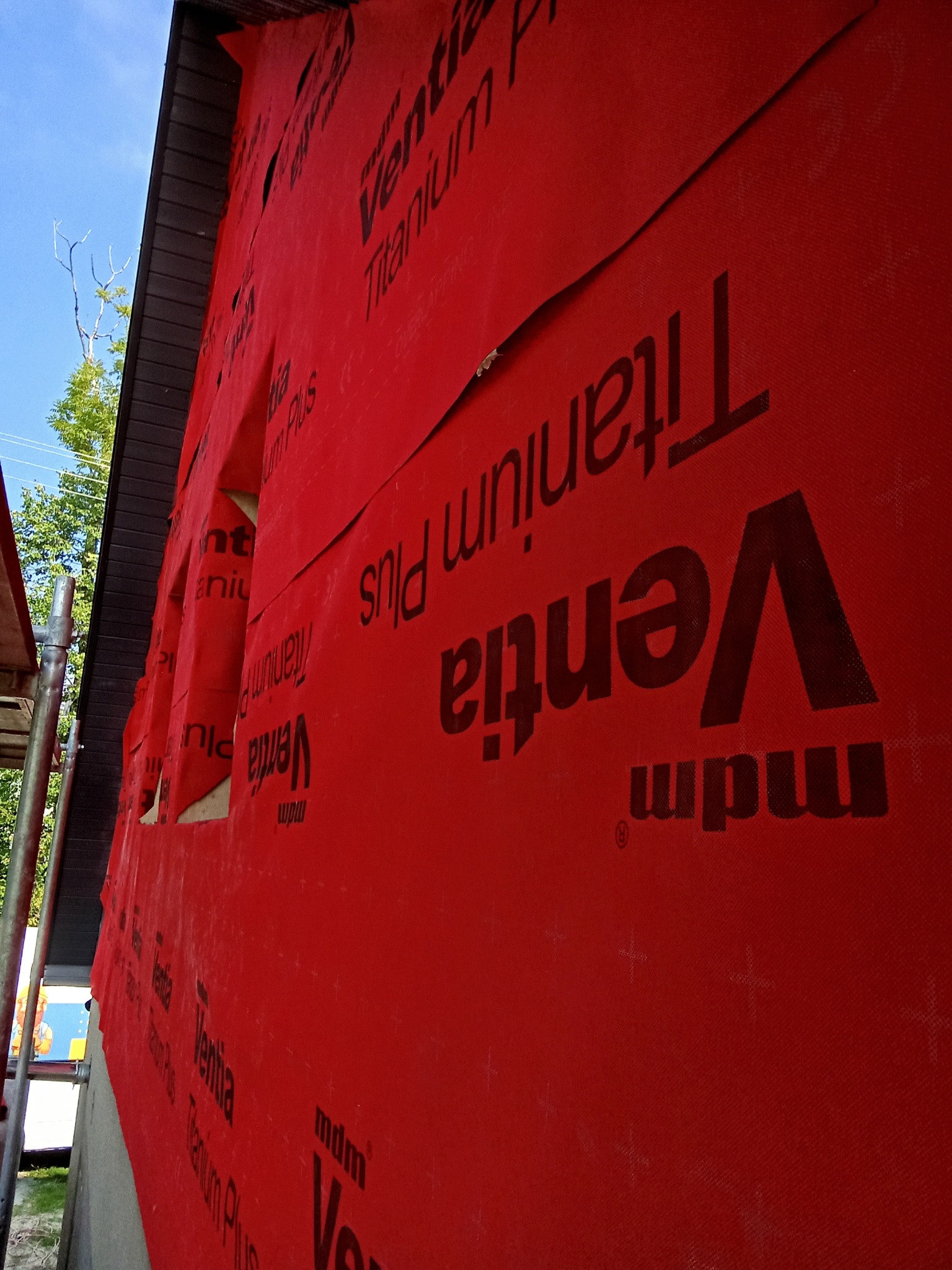 Czerwona membrana wiatroizolacyjna z czarnym nadrukiem 'Ventia Titanium Plus' na ścianie budynku w trakcie budowy, widoczne otwory okienne i fragment rusztowania.