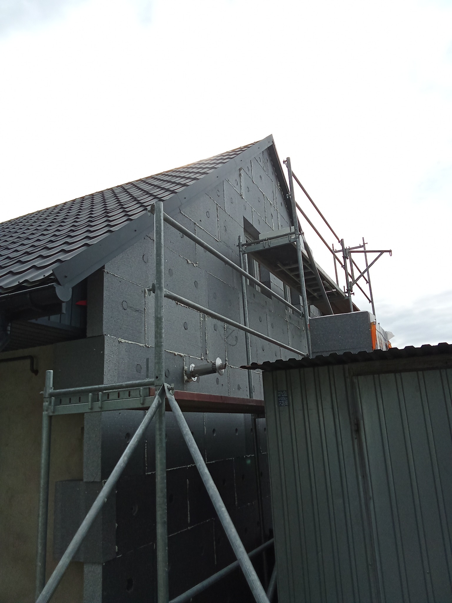Ocieplanie domu styropianem grafitowym, widoczne rusztowanie i fragment dachu z blachodachówki. Prace w toku, widok z dołu na elewację budynku.