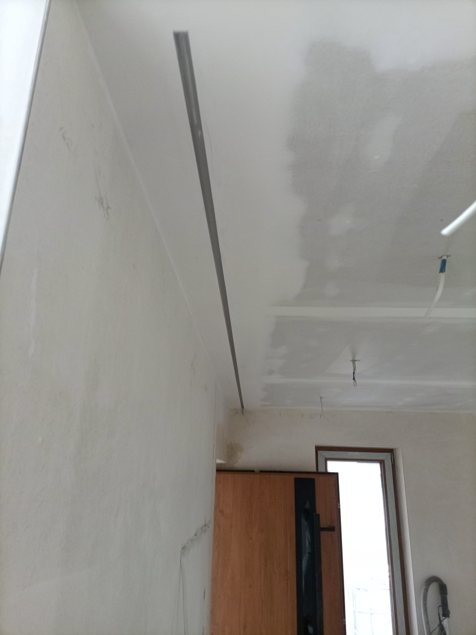 Wnętrze w trakcie prac wykończeniowych: ściana z widocznymi śladami po gładzi, zamontowany profil sufitowy i częściowo wykończony sufit. Widoczne kable i drzwi w tle.