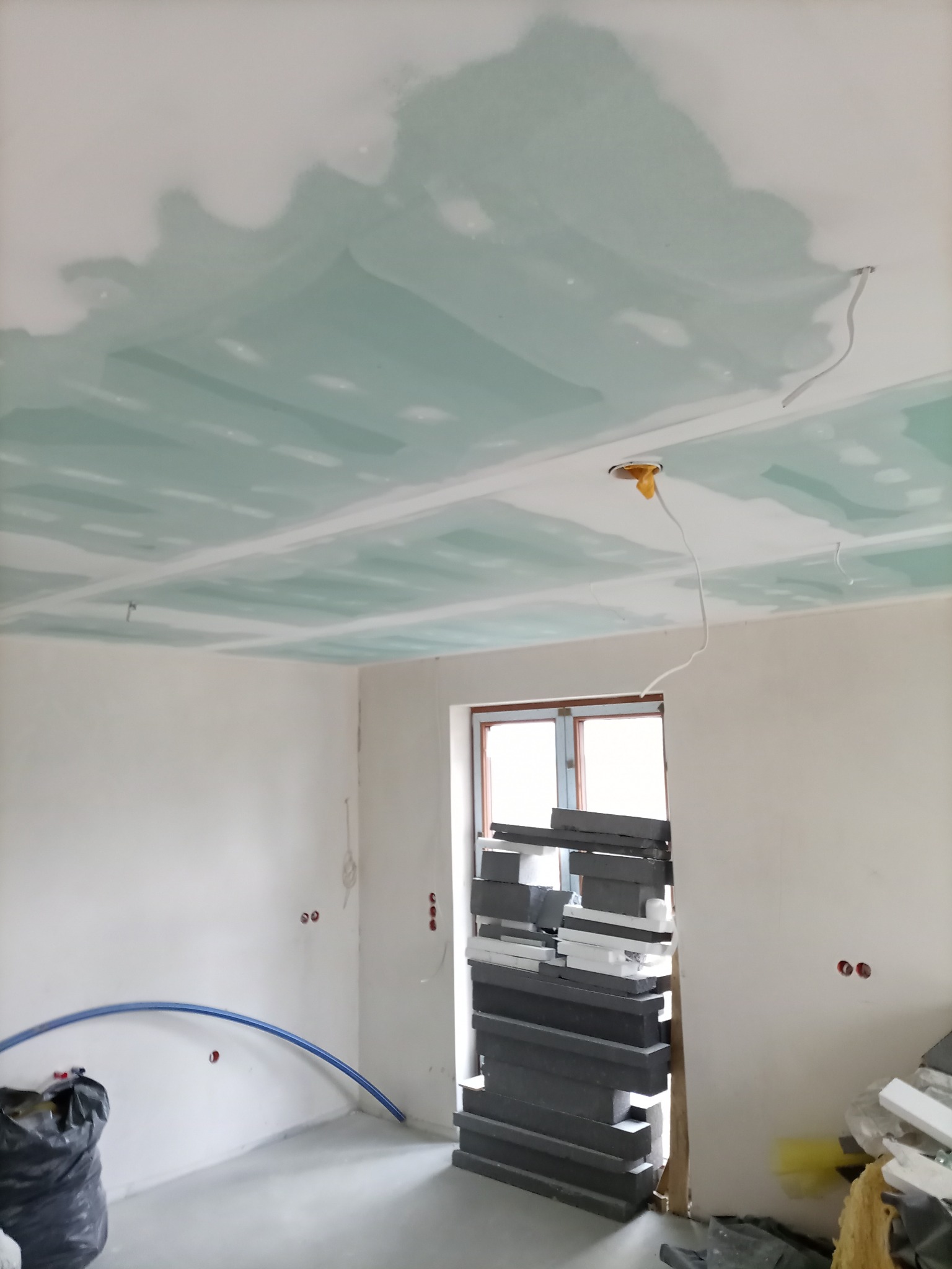 Wnętrze w trakcie prac wykończeniowych: sufit z płyt karton-gips, widoczne łączenia, instalacja elektryczna, materiały budowlane przy oknie. Ściany przygotowane do malowania.