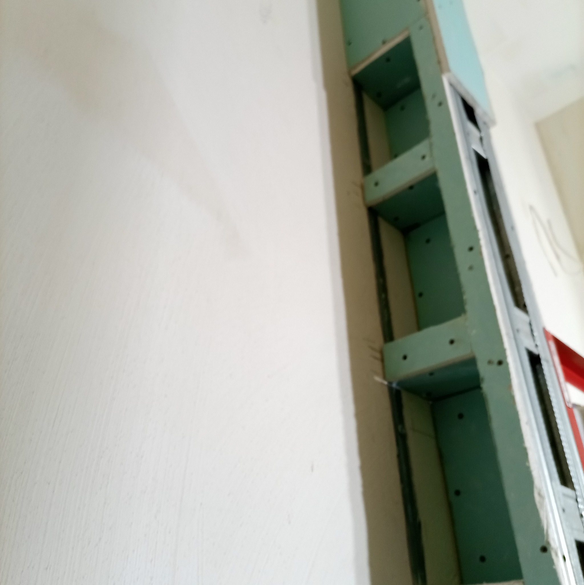 Narożnik ściany z konstrukcją z profili i zielonej płyty GK, tworzącą półki wnękowe. Widoczna surowa ściana obok. Ujęcie z dołu, podkreślające pionowość konstrukcji.