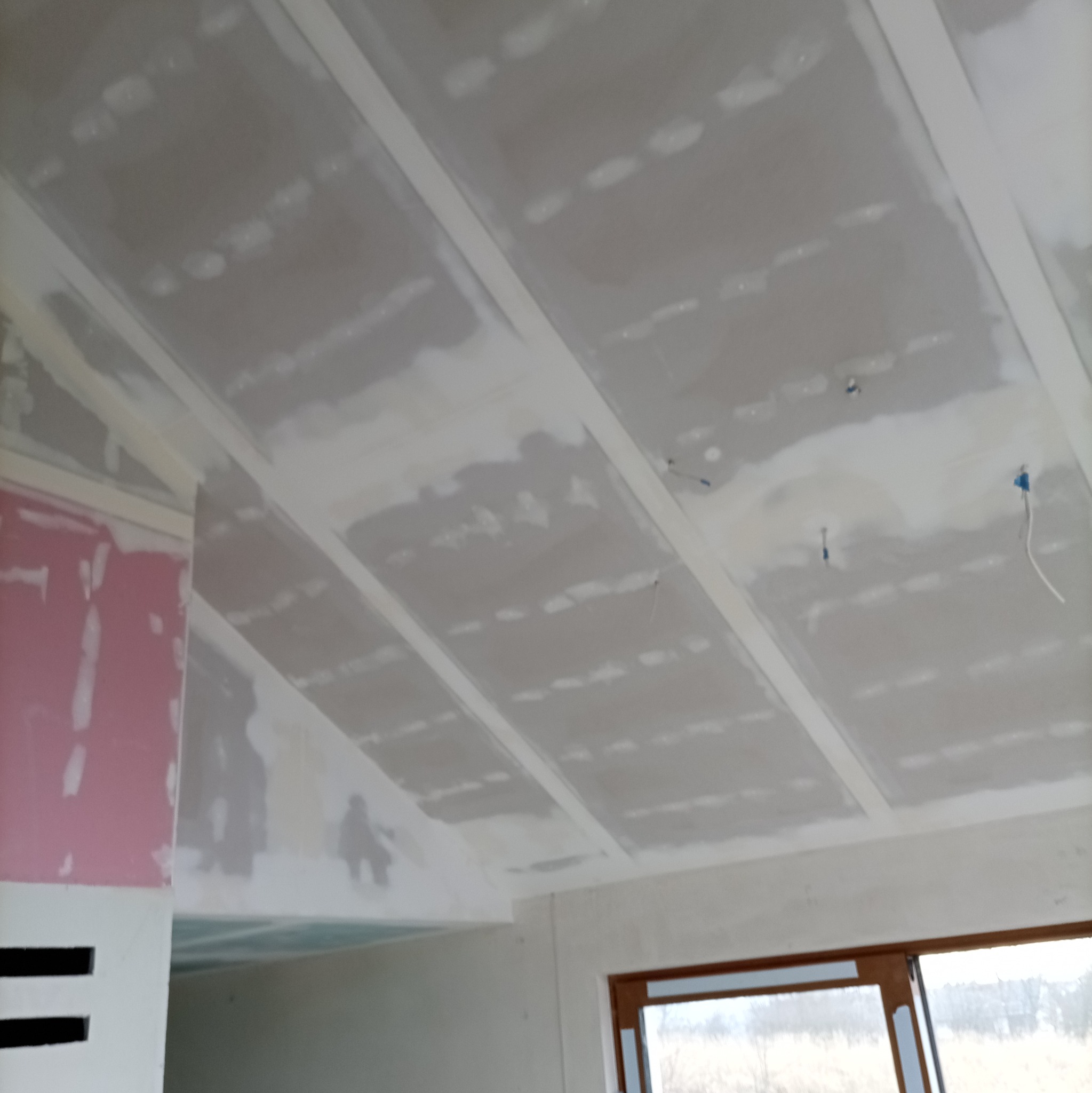 Skośny sufit poddasza z płyt kartonowo-gipsowych, częściowo zaszpachlowany, widoczne profile i kable. Okno w tle.