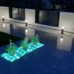 Nocne ujęcie tarasu z ciemnymi płytkami, podświetlaną rabatą z turkusowymi kamieniami i roślinami, białymi kamykami przy ogrodzeniu z poziomymi panelami oraz oświetleniem punktowym.