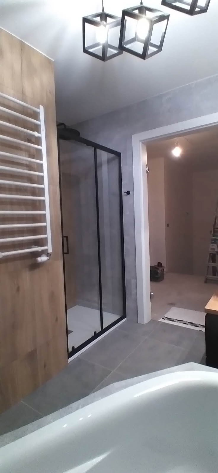 Nowoczesna łazienka w trakcie wykończenia, widoczna kabina prysznicowa z czarnymi profilami, grzejnik drabinkowy na drewnopodobnej ścianie, szare płytki podłogowe i fragment wanny na pierwszym...