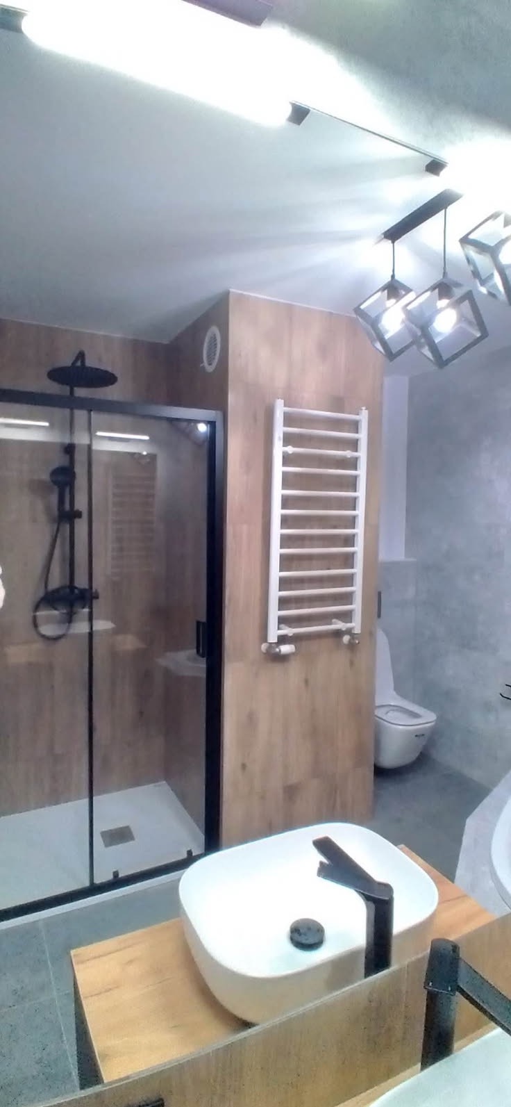 Nowoczesna łazienka z kabiną prysznicową typu walk-in z czarnymi ramami, białą grzejnikiem drabinkowym, umywalką nablatową na drewnianym blacie i geometryczną lampą sufitową.
