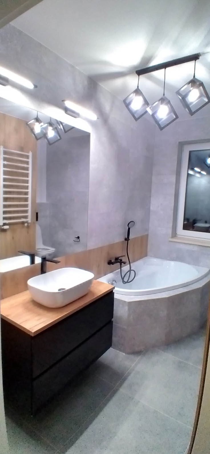 Nowoczesna łazienka z umywalką nablatową na szafce z drewnianym blatem, narożną wanną obudowaną szarą mozaiką oraz designerską lampą sufitową z czterema kwadratowymi kloszami.
