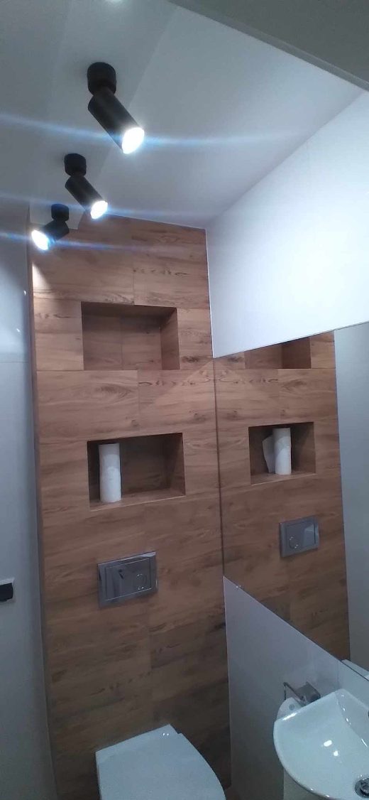 Nowoczesna łazienka z drewnopodobnymi płytkami, wnękami w ścianie podświetlonymi trzema reflektorami sufitowymi, toaleta i umywalka widoczne w odbiciu lustra.