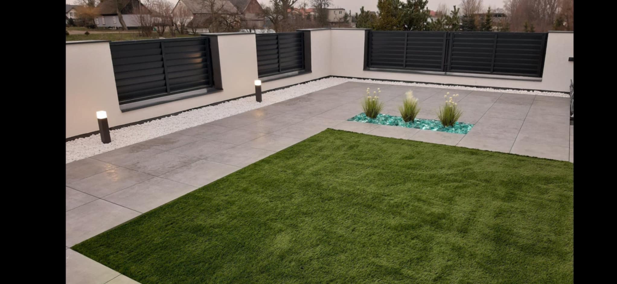 Nowoczesny taras z szarych płyt, sztuczną trawą, białym żwirem, podświetlanym ogrodzeniem i dekoracyjnymi roślinami w turkusowym szkle.