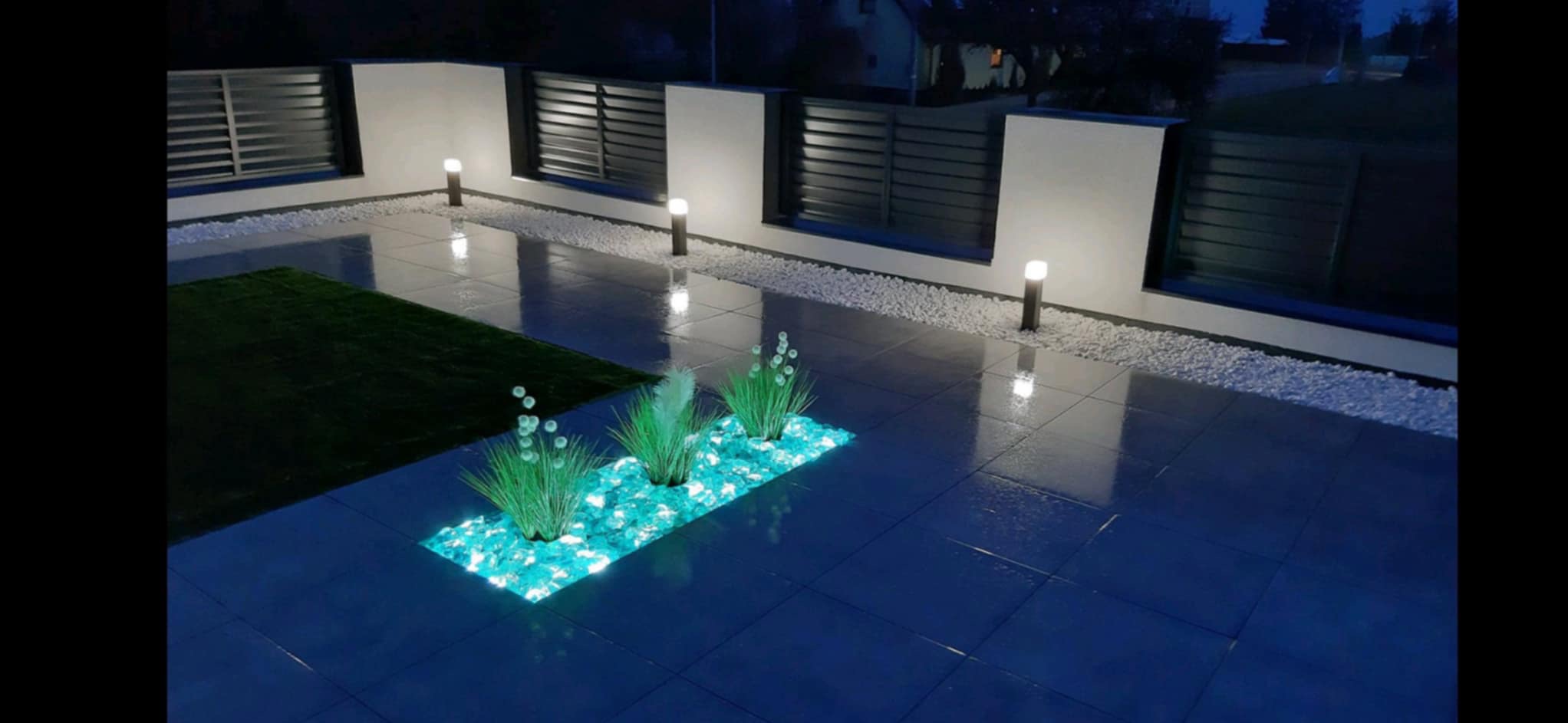 Nocne ujęcie tarasu z ciemnymi płytkami, podświetlaną rabatą z turkusowymi kamieniami i roślinami, białymi kamykami przy ogrodzeniu z poziomymi panelami oraz oświetleniem punktowym.