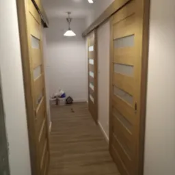 Korytarz z drewnianymi drzwiami przesuwnymi z przeszkleniami, widoczne dwa źródła światła na suficie i podłoga imitująca drewno.