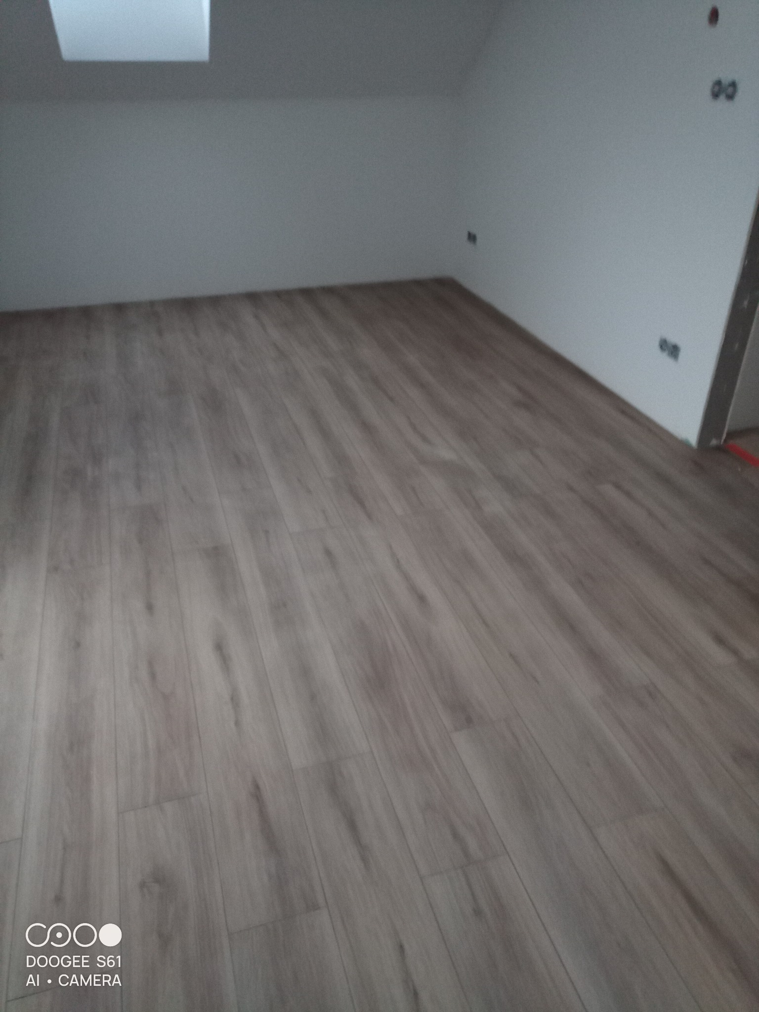 Ukończona podłoga z paneli w pokoju na poddaszu, widoczne gniazdka elektryczne na ścianie i okno dachowe.