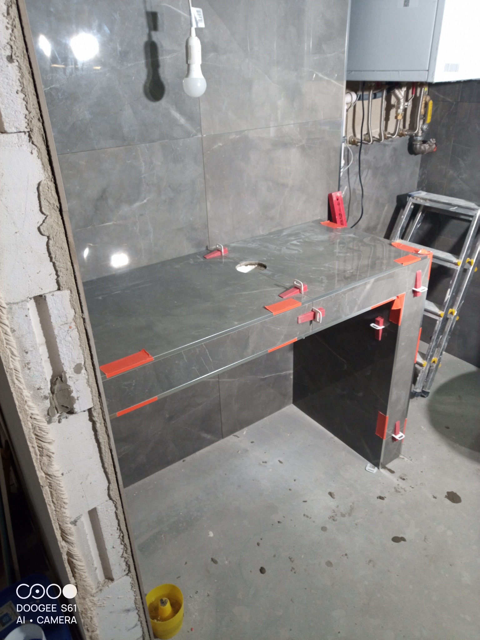 Montaż szarej, marmurkowej umywalki z płytek, z użyciem czerwonych klinów poziomujących, widoczna drabina i fragment instalacji CO.