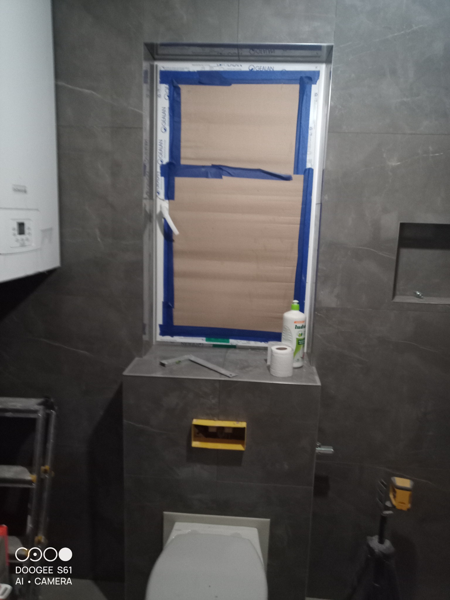 Remont łazienki w trakcie: szare płytki, zamaskowane okno taśmą malarską, toaleta, bojler, poziomica i laser krzyżowy.