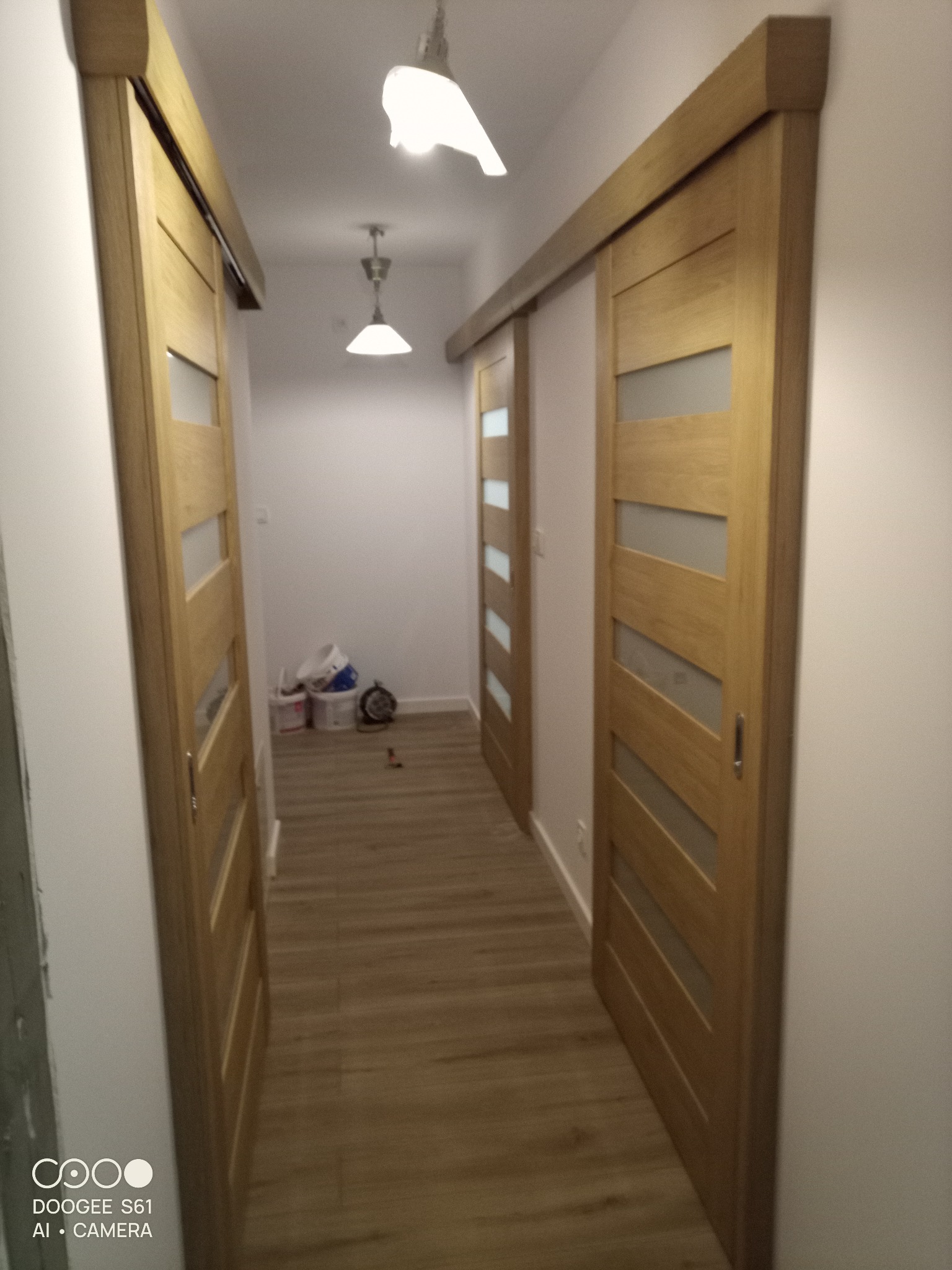 Korytarz z drewnianymi drzwiami przesuwnymi z przeszkleniami, widoczne dwa źródła światła na suficie i podłoga imitująca drewno.