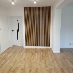 LUX - REM ŁUKASZ NIEDBALSKI - Jasne panele podłogowe w nowocześnie wykończonym wnętrzu z drewnianą ścianką dekoracyjną i białą stolarką drzwiową z czarnym, falistym akcentem.