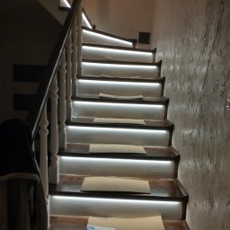 LUX - REM ŁUKASZ NIEDBALSKI - Oświetlone schody drewniane z białymi poręczami i dekoracyjną ścianą. Podświetlenie LED pod każdym stopniem. Widok z dołu na górę.