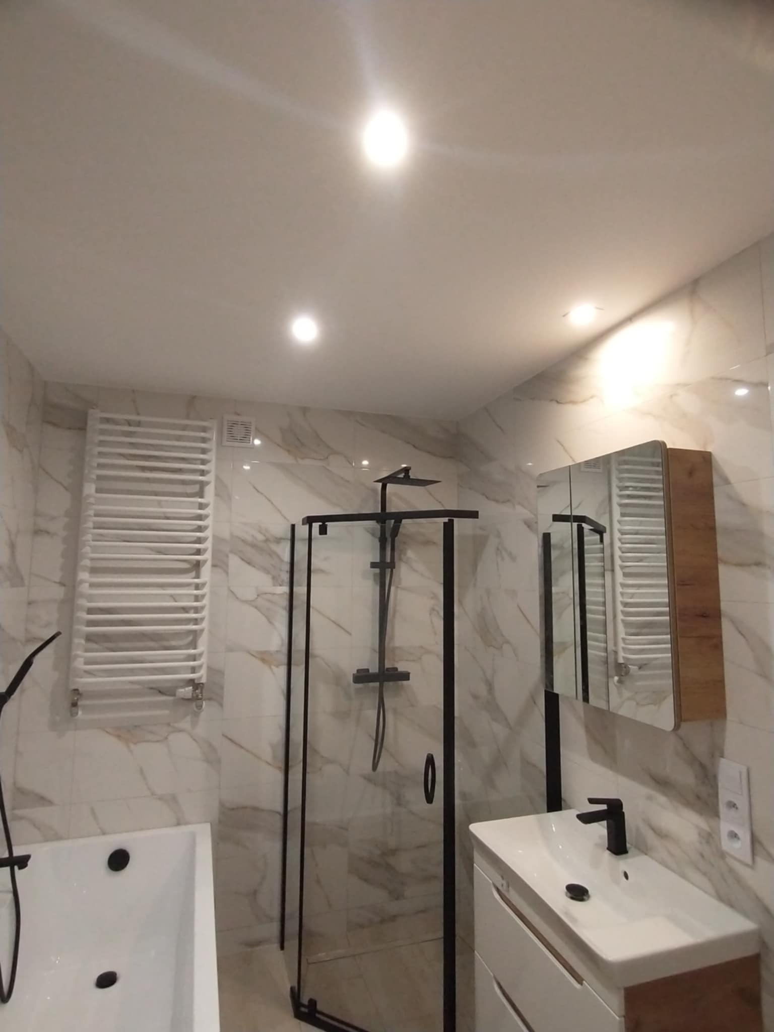 Wykończona łazienka z kabiną prysznicową typu walk-in, białą wanną, grzejnikiem drabinkowym i szafką z lustrem. Marmurowe płytki na ścianach i sufitowe oświetlenie punktowe.