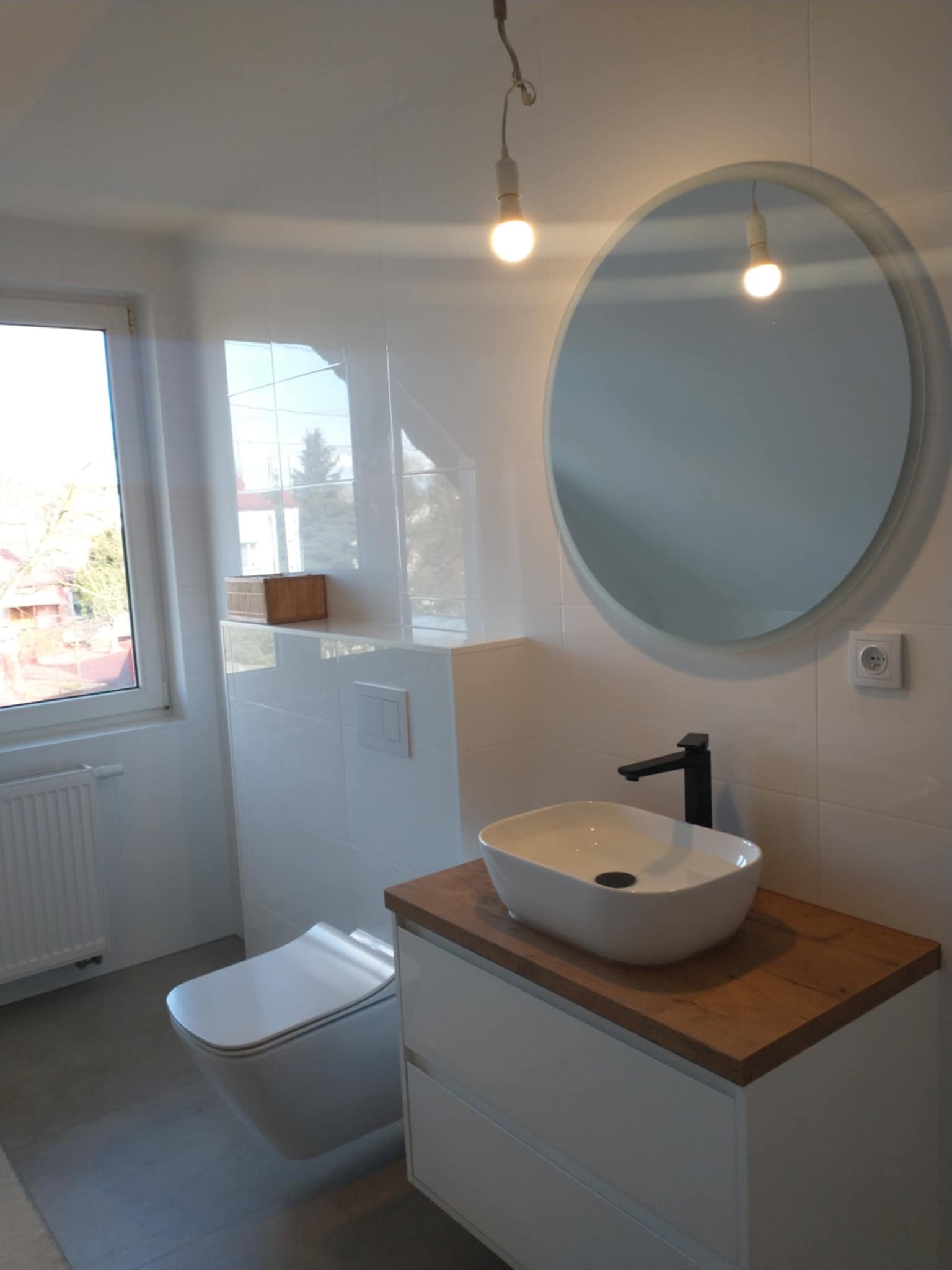 Nowoczesna łazienka z owalnym lustrem, białą umywalką na drewnianym blacie i podwieszaną toaletą. Białe płytki na ścianach, minimalistyczny design.