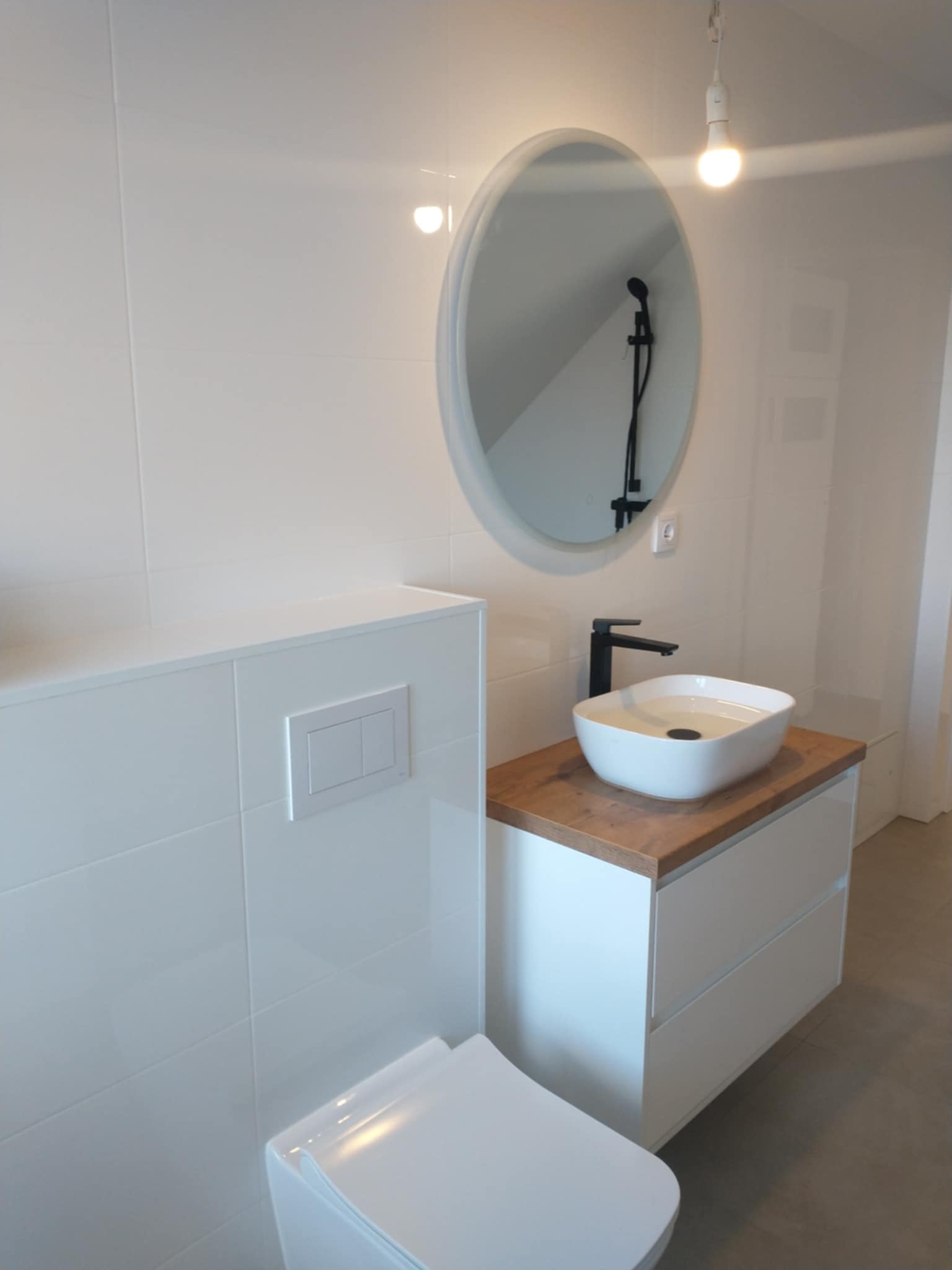Nowoczesna łazienka z owalnym lustrem, białą umywalką na drewnianym blacie i podwieszaną toaletą. Minimalistyczny design, białe płytki i czarna armatura.
