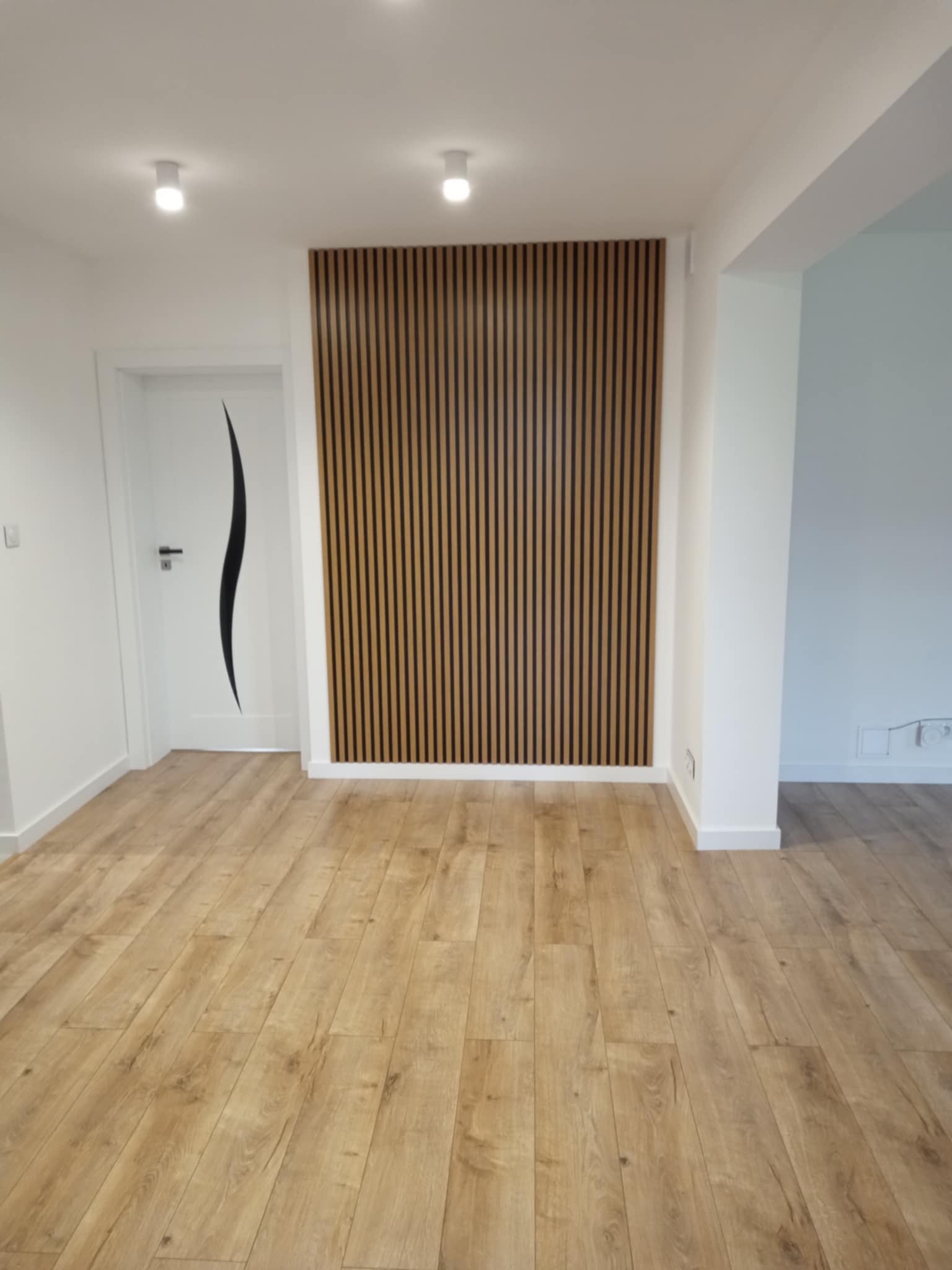 Jasne panele podłogowe w nowocześnie wykończonym wnętrzu z drewnianą ścianką dekoracyjną i białą stolarką drzwiową z czarnym, falistym akcentem.