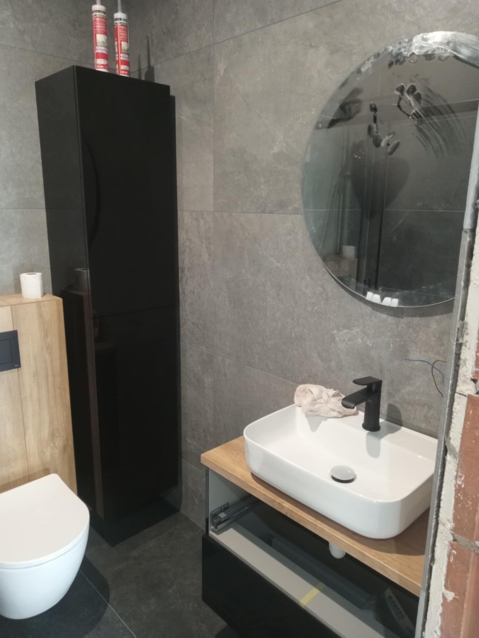 Nowoczesna łazienka z szarą płytką, okrągłym lustrem z zaciekami, białą umywalką na drewnianym blacie i czarną szafką. Widoczne silikony na szafce.