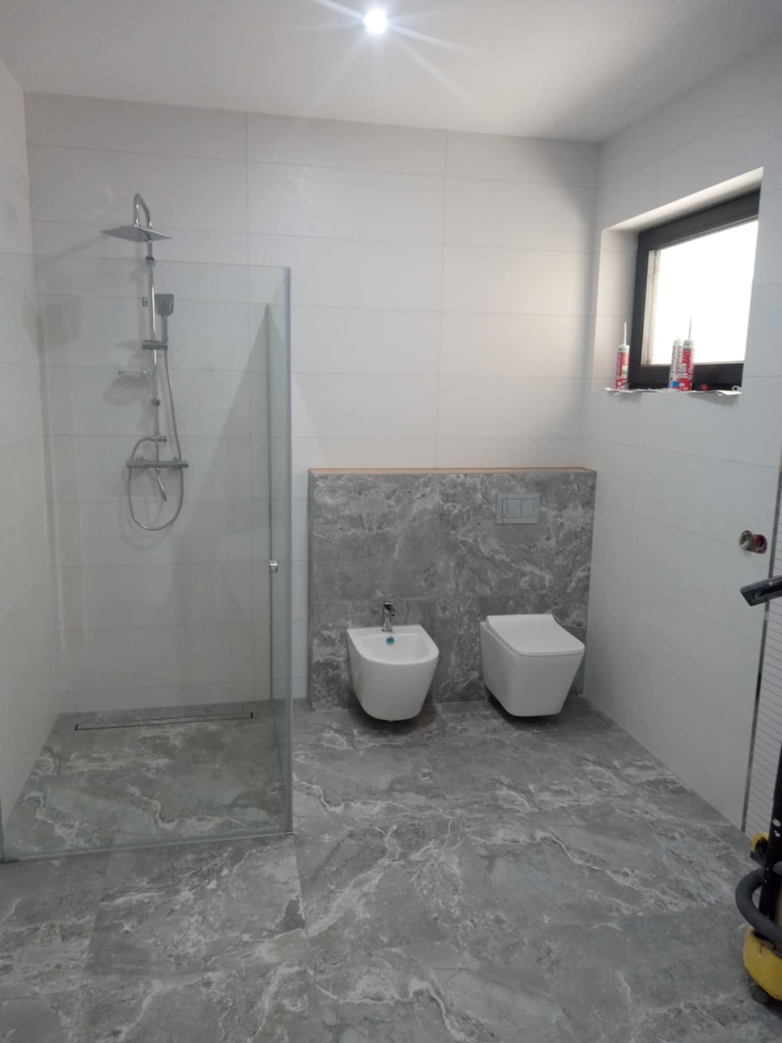 Wykończona łazienka z kabiną prysznicową typu walk-in, bidetem i toaletą w stylu nowoczesnym. Szare płytki imitujące marmur na podłodze i ścianie.