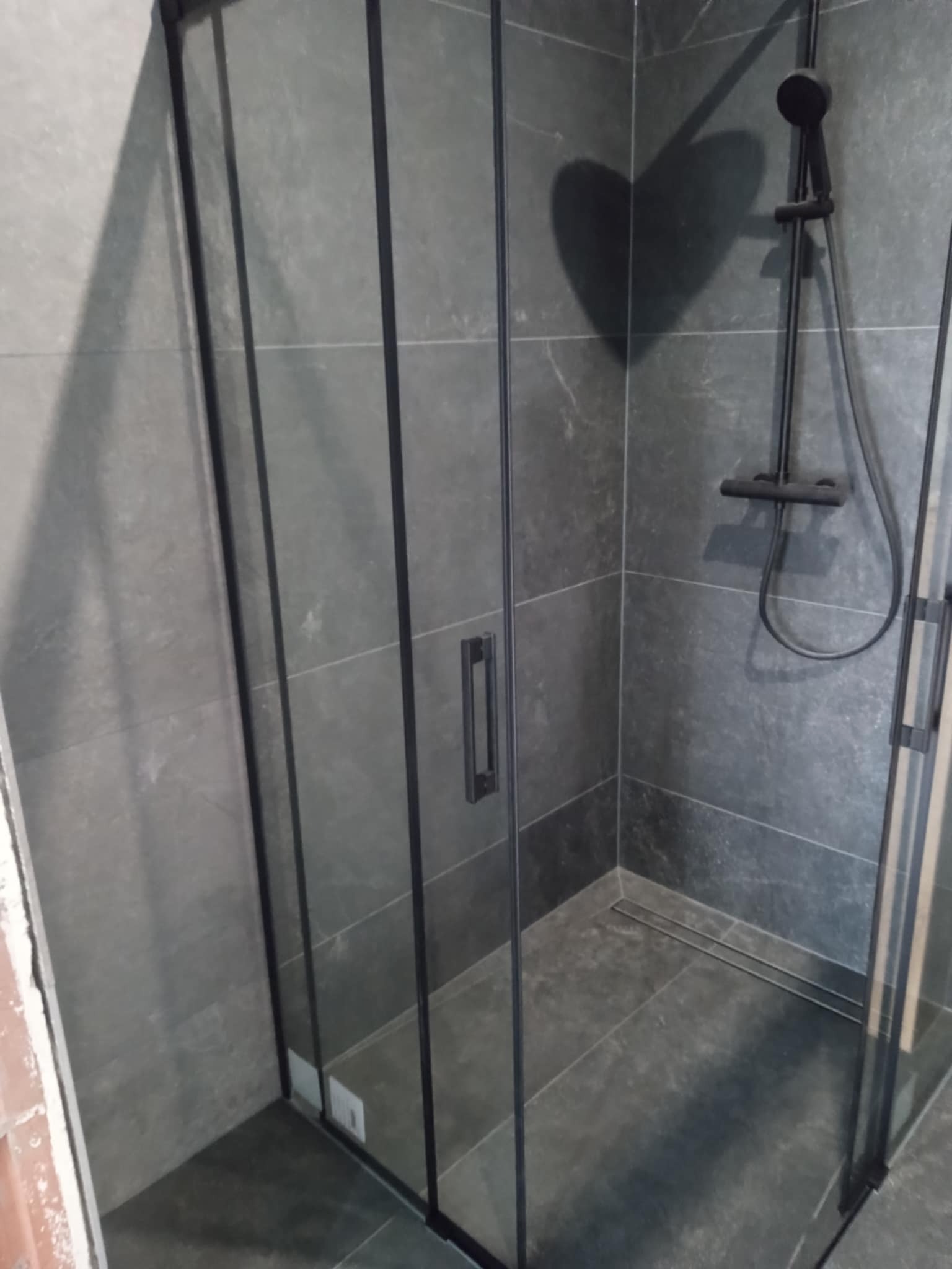 Nowoczesna kabina prysznicowa z czarnymi profilami i szarymi płytkami. Widoczny odpływ liniowy i deszczownica. Minimalistyczny design łazienki.