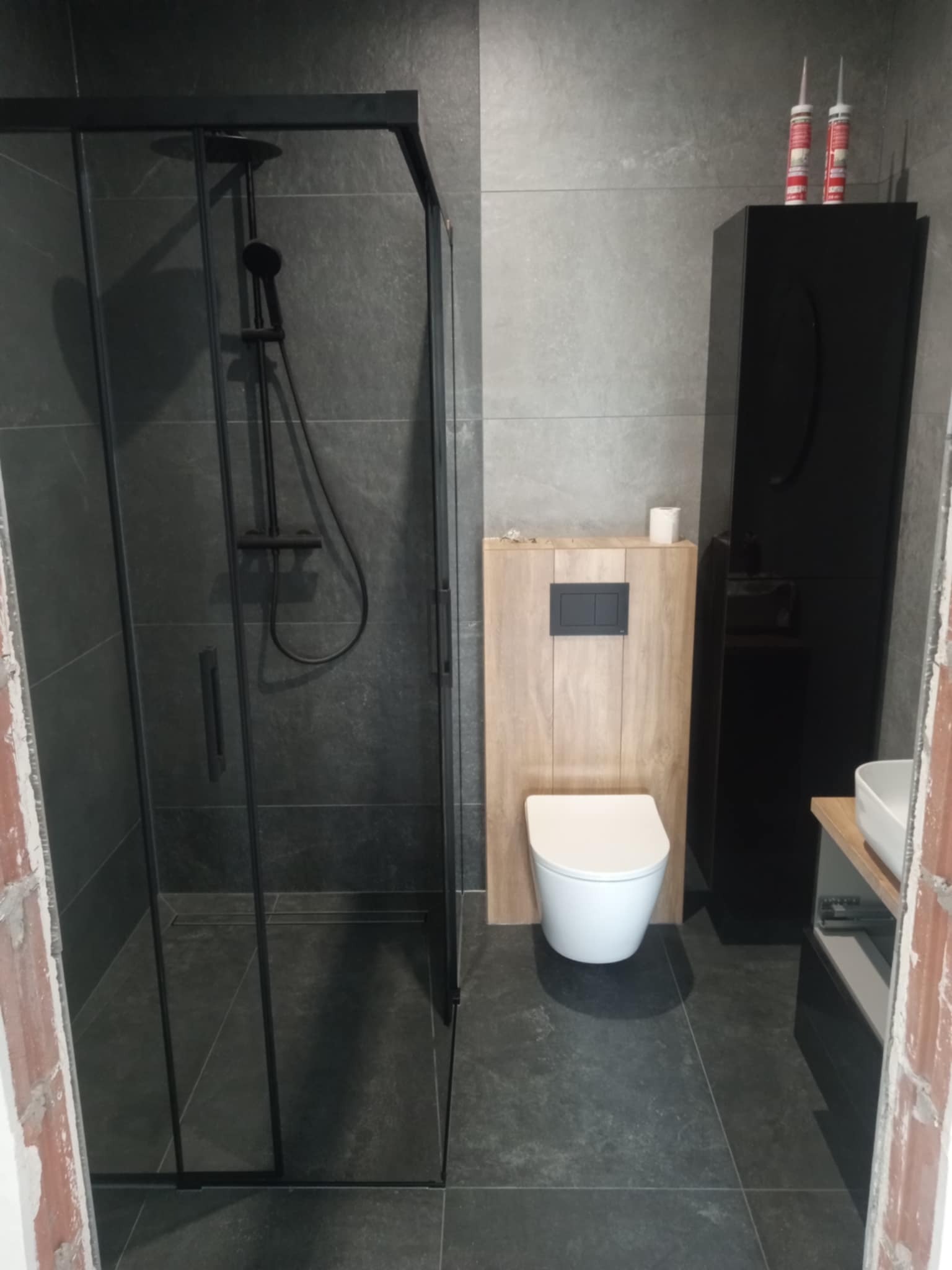 Nowoczesna łazienka w Osjakowie z kabiną prysznicową typu walk-in, wiszącą toaletą i czarną szafką. Szare płytki i minimalistyczny design.