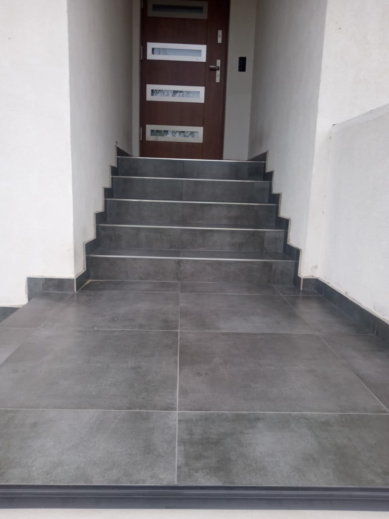 Schody wejściowe wyłożone szarymi płytkami, z widocznymi fugami i ciemną listwą przypodłogową. Brązowe drzwi z przeszkleniami w tle. Minimalistyczny design.