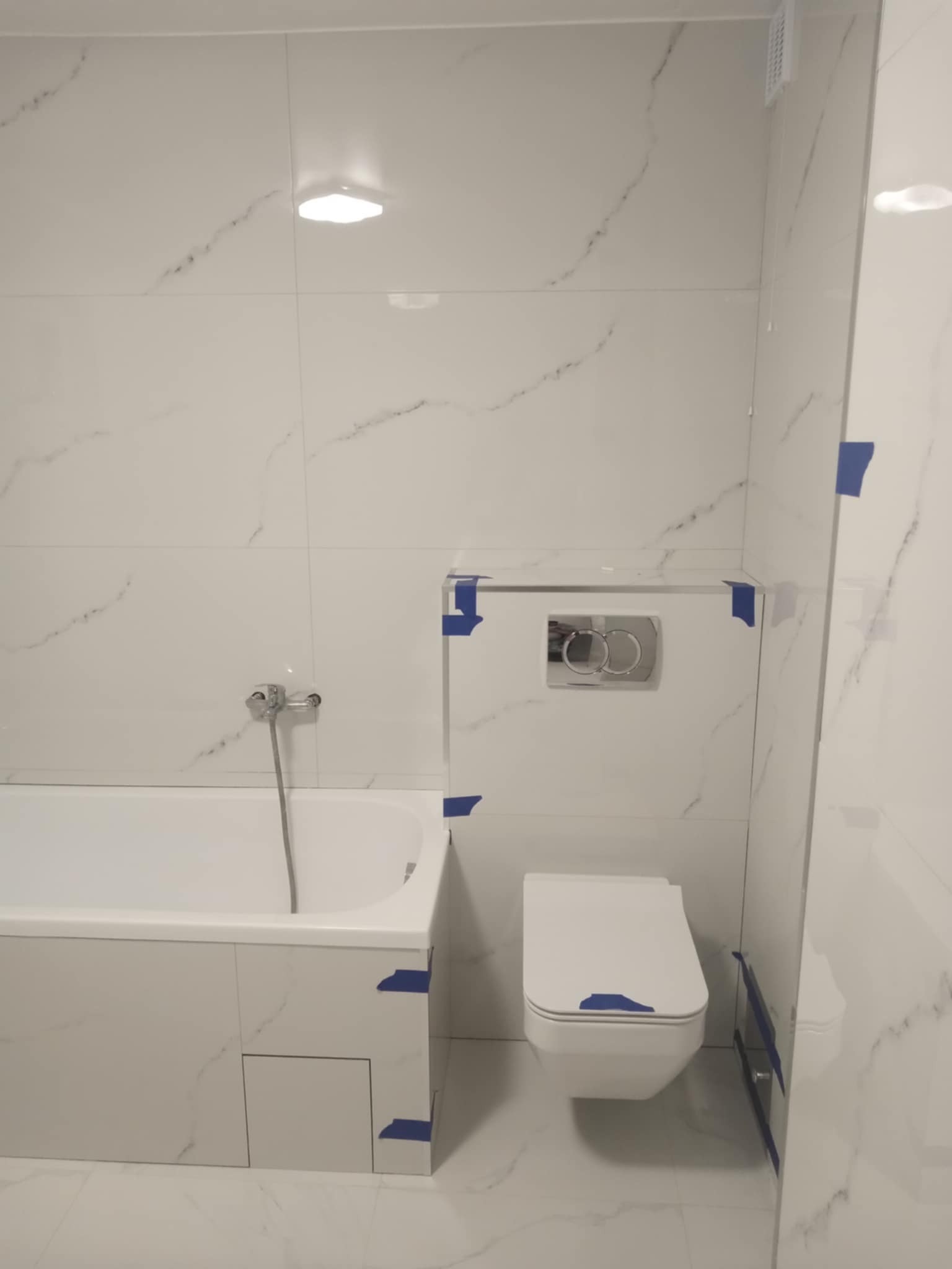 Biała łazienka z wanną, toaletą podwieszaną i płytkami imitującymi marmur. Zabezpieczenia taśmą malarską na łączeniach. Minimalistyczny design, jasne oświetlenie.