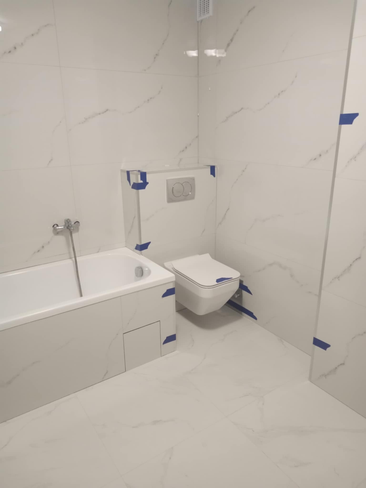 Wykończona łazienka w bieli z marmurowym wzorem płytek, widoczna wanna, toaleta podwieszana i fragment ściany z niebieską taśmą zabezpieczającą.