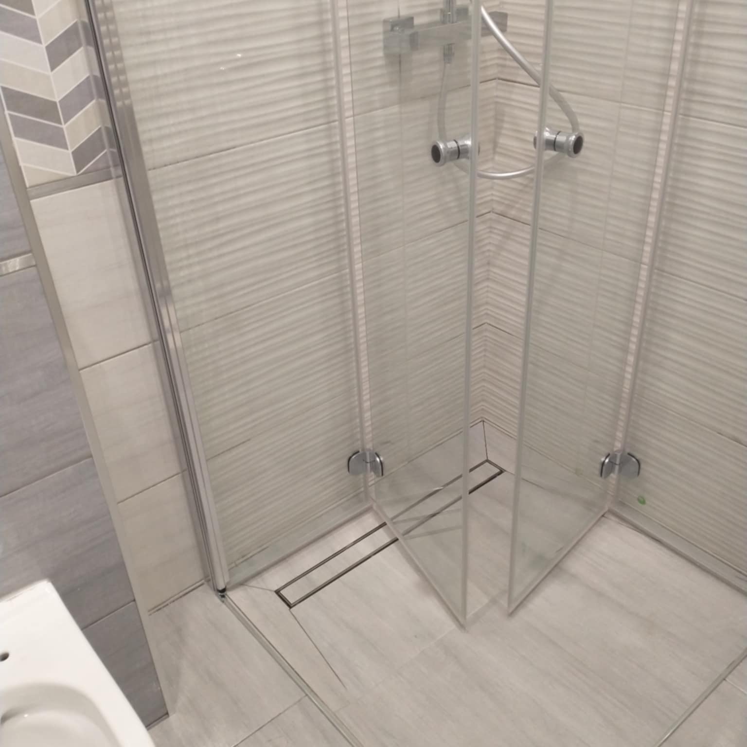 Nowoczesna kabina prysznicowa z odpływem liniowym, wykończona płytkami w odcieniach szarości i bieli. Widoczny fragment umywalki. Minimalistyczny design.