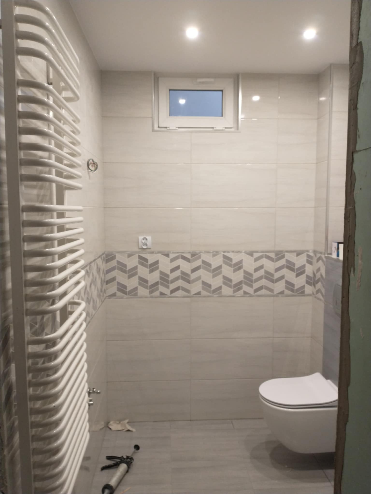 Wykończona łazienka z białymi płytkami, dekoracyjnym pasem z szarymi wzorami, wiszącą toaletą i grzejnikiem drabinkowym. Widoczne narzędzia glazurnicze.