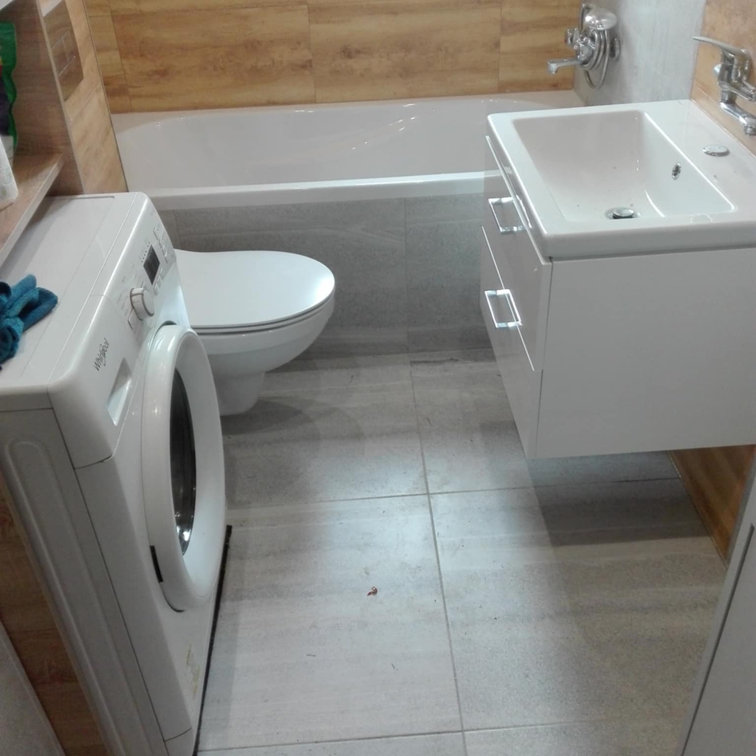 Wykończona łazienka z pralką, umywalką z szafką i wanną w jasnych odcieniach szarości i beżu. Widoczne płytki na podłodze i ścianach.