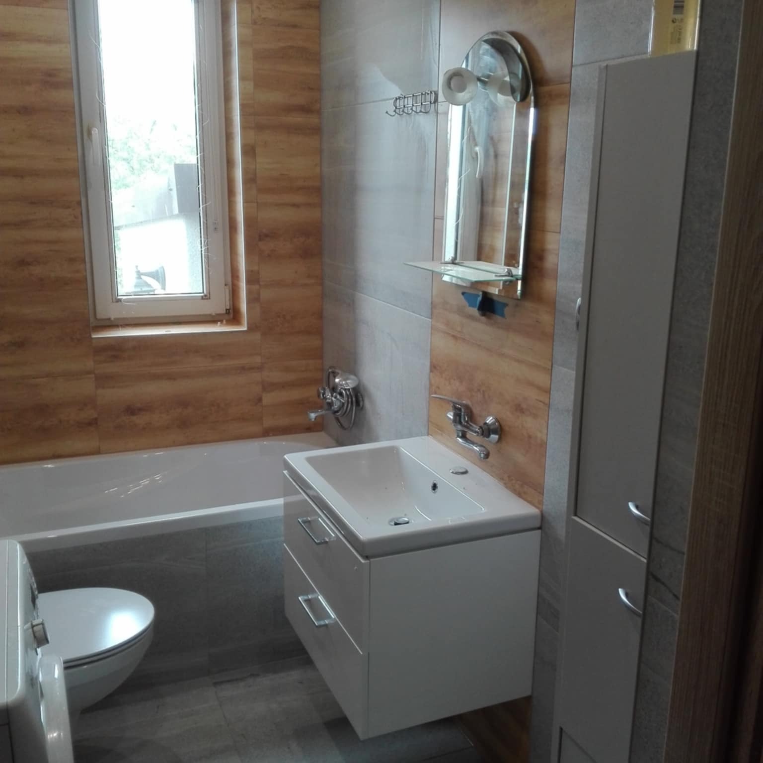 Nowoczesna łazienka z drewnopodobnymi płytkami, białą armaturą i szafką pod umywalką z szufladami. Lustro z półką i relingiem na ręczniki. Okno z widokiem na zewnątrz.