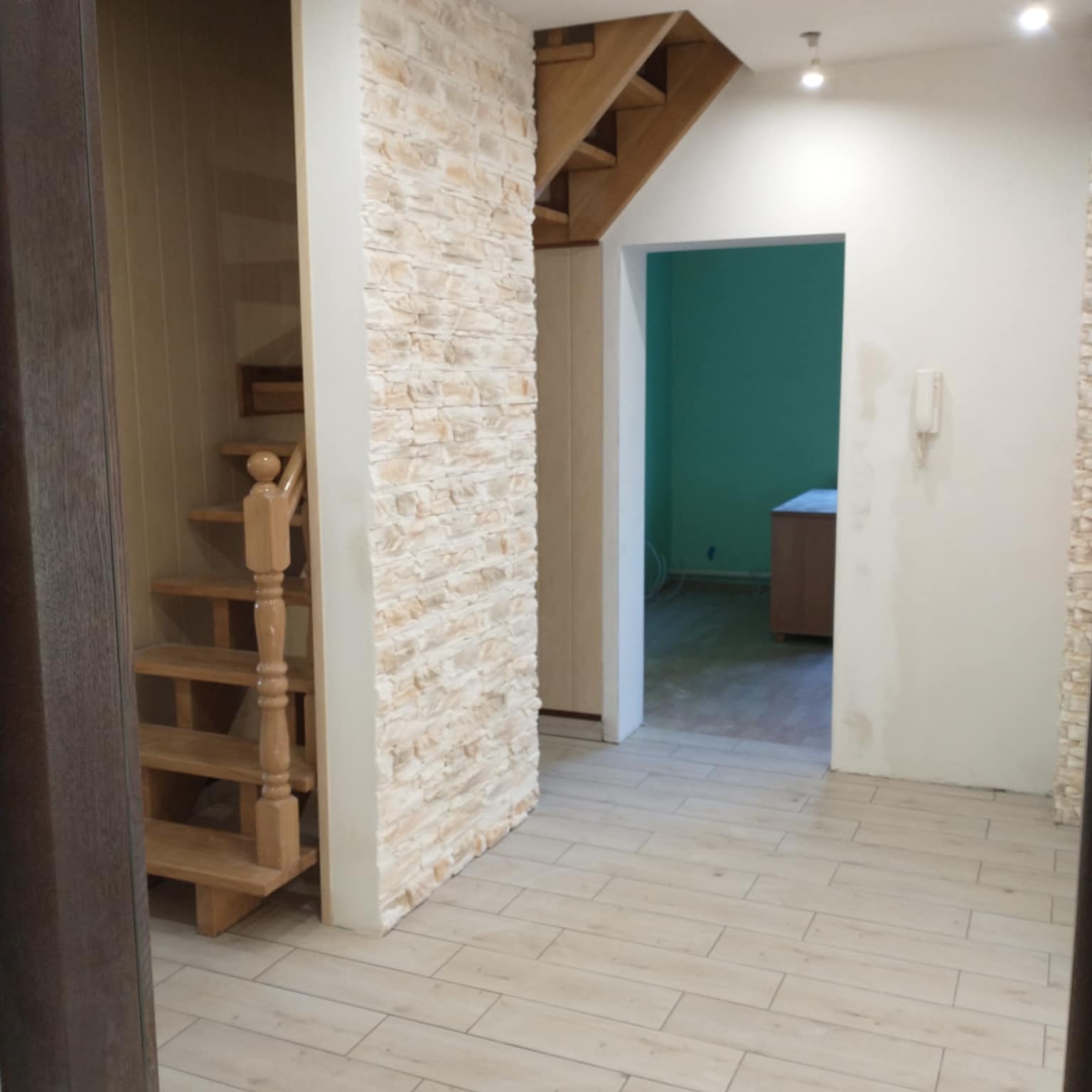 Wykończone wnętrze z drewnianymi schodami i dekoracyjną ścianą z kamienia. Jasna podłoga z paneli imitujących drewno. Widok na pomalowane na zielono pomieszczenie.