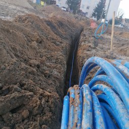 Patryk Majkut - Wykopany rów w ziemi z ułożonym niebieskim przewodem instalacyjnym, widoczne zwoje przewodu na pierwszym planie, w tle budynek mieszkalny.
