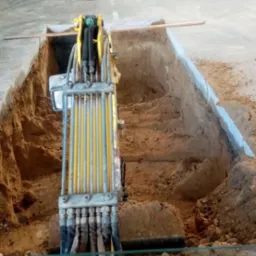 Żółta maszyna kopiąca wąski wykop wewnątrz hali przemysłowej, widoczna z perspektywy operatora, w tle zaparkowany samochód terenowy i palety z deskami.